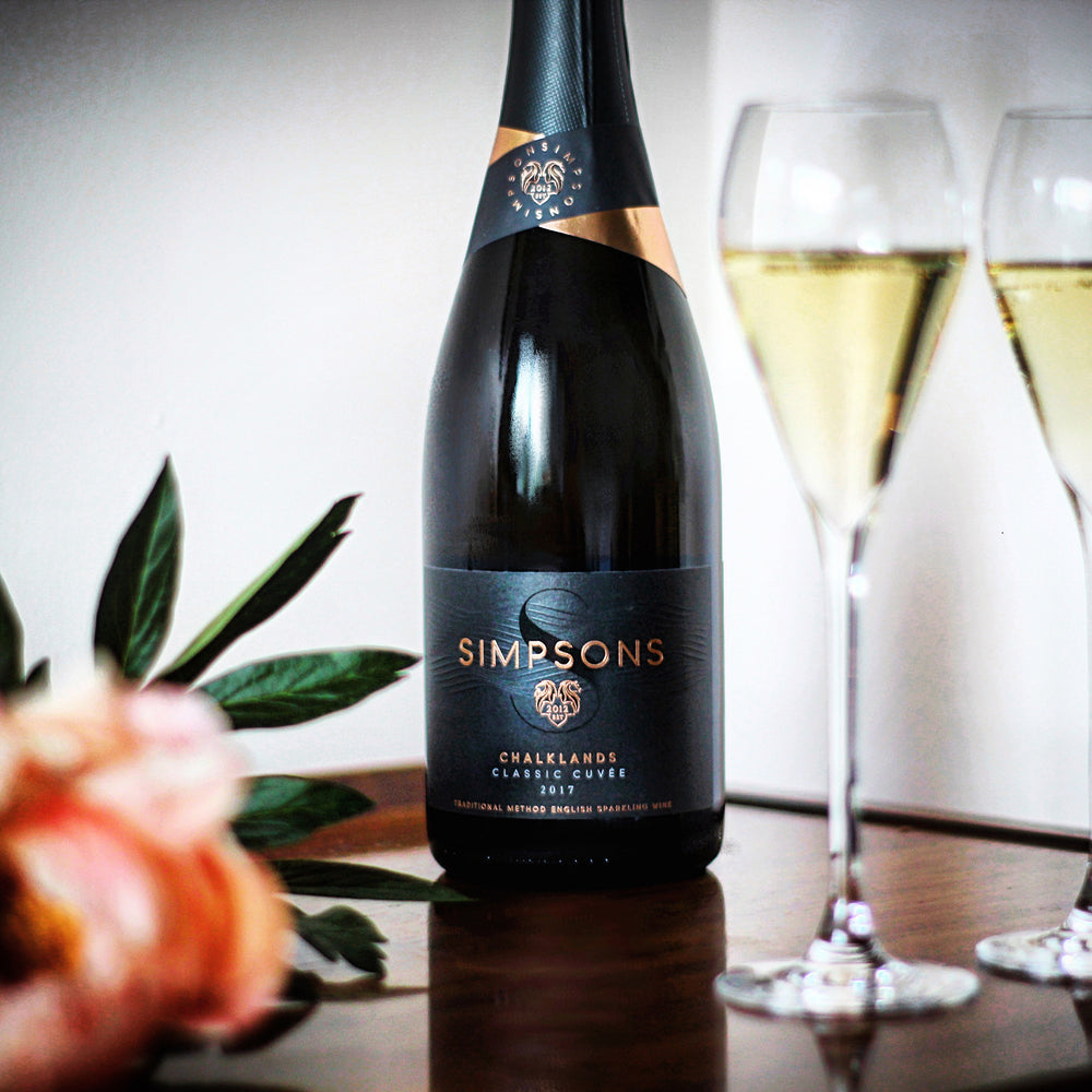 A bottle of Simpsons sparkling wine on a table next to a flower and two glasses