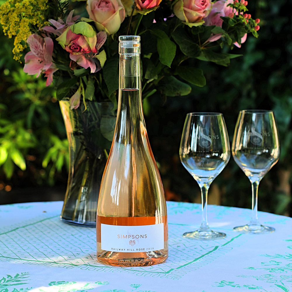 A bottle of Simpsons still wine on a garden table with a vase of flowers and two wine glasses